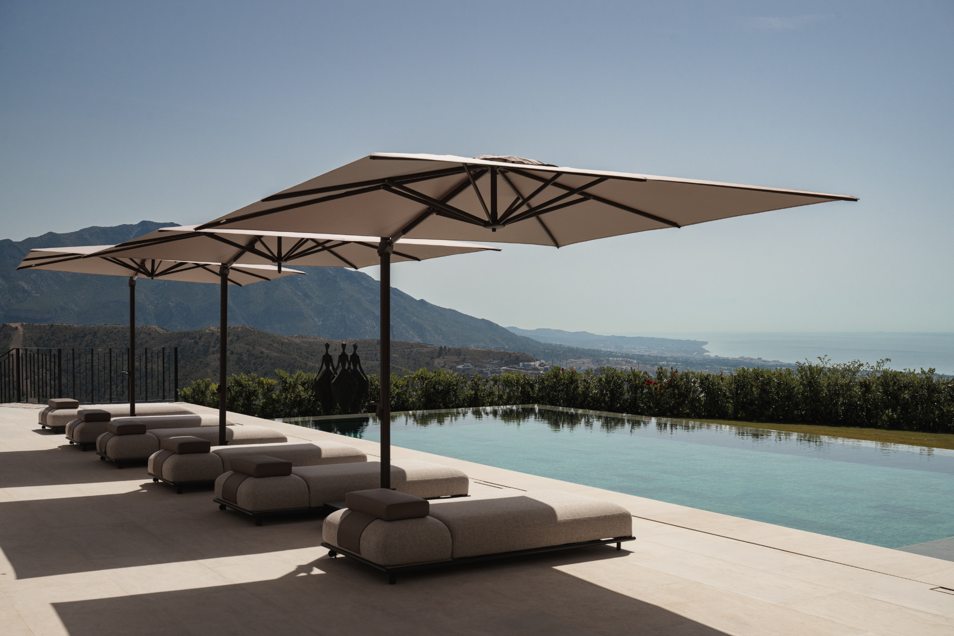 Loungers beneath cantilevered parasols beside the infinity pool, looking out over the mountains toward the Mediterranean coast.